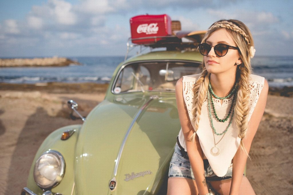 Imagem com fundo de uma praia e o mar azul. Em destaque a imagem de um fusca vintage na cor verde claro. Sobre ele, um caixa vermelha com o logo da marca Coca Cola, escrito em branco e uma prancha de surfe. À frente do carro a figura de uma mulher estilo hippie, loura, com cabelos longos com duas tranças. Ela usa óculos escuros, colares, uma blusa branca, shorts jeans e uma flor na cabeça.