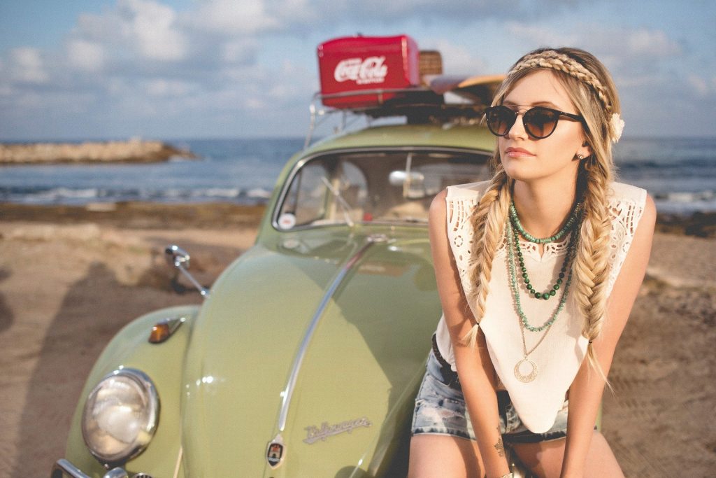 Imagem com fundo de uma praia e o mar azul. Em destaque a imagem de um fusca vintage na cor verde claro. Sobre ele, um caixa vermelha com o logo da marca Coca Cola, escrito em branco e uma prancha de surfe. À frente do carro a figura de uma mulher estilo hippie, loura, com cabelos longos com duas tranças. Ela usa óculos escuros, colares, uma blusa branca, shorts jeans e uma flor na cabeça.
