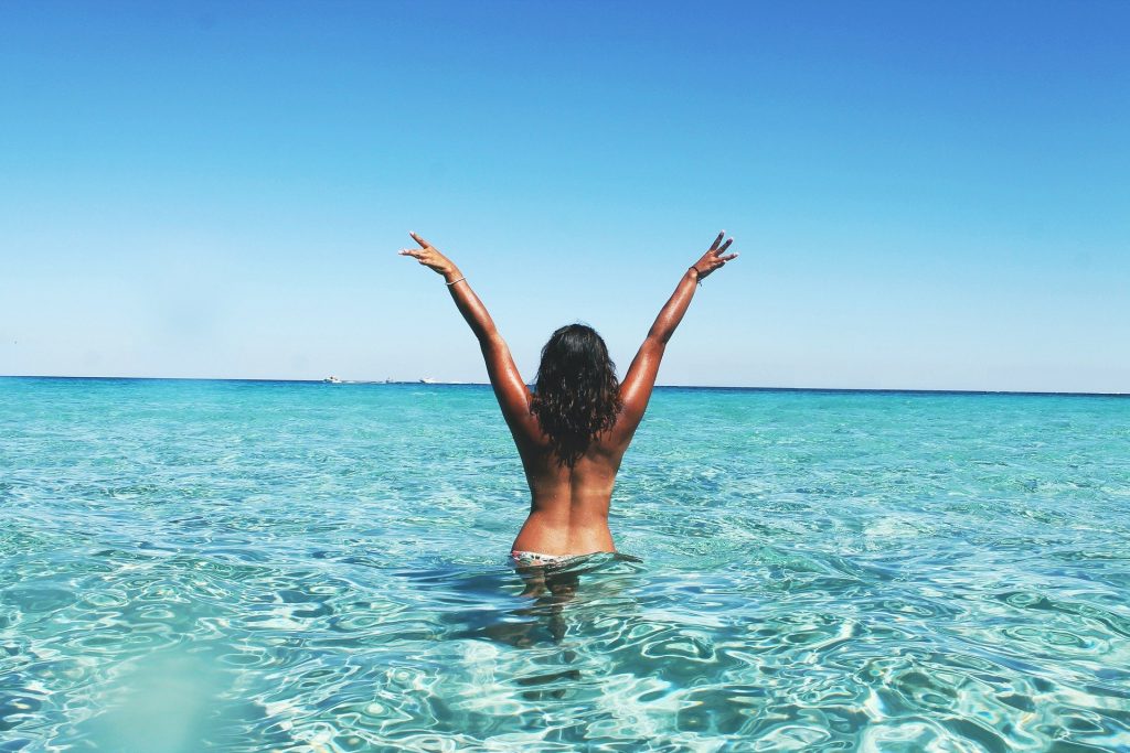 Imagem de um lindo mar azul e nele uma mulher de costas com os braços levantados. Ela está bronzeada e em busca de liberdade.