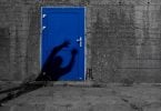 Imagem de uma parede cinza com a entrada para uma porta pintada na cor azul bic. Na porta consta a sombra de um fantasma que está querendo assombrar alguém.