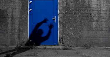 Imagem de uma parede cinza com a entrada para uma porta pintada na cor azul bic. Na porta consta a sombra de um fantasma que está querendo assombrar alguém.