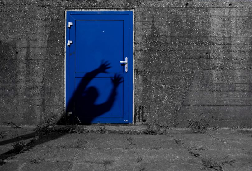 Imagem de uma parede cinza com a entrada para uma porta pintada na cor azul bic. Na porta consta a sombra de um fantasma que está querendo assombrar alguém.