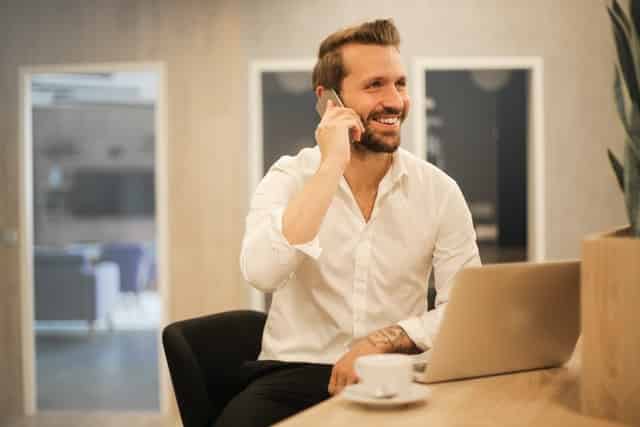 Homem branco sentado com celular no ouvido e notebook na mesa.