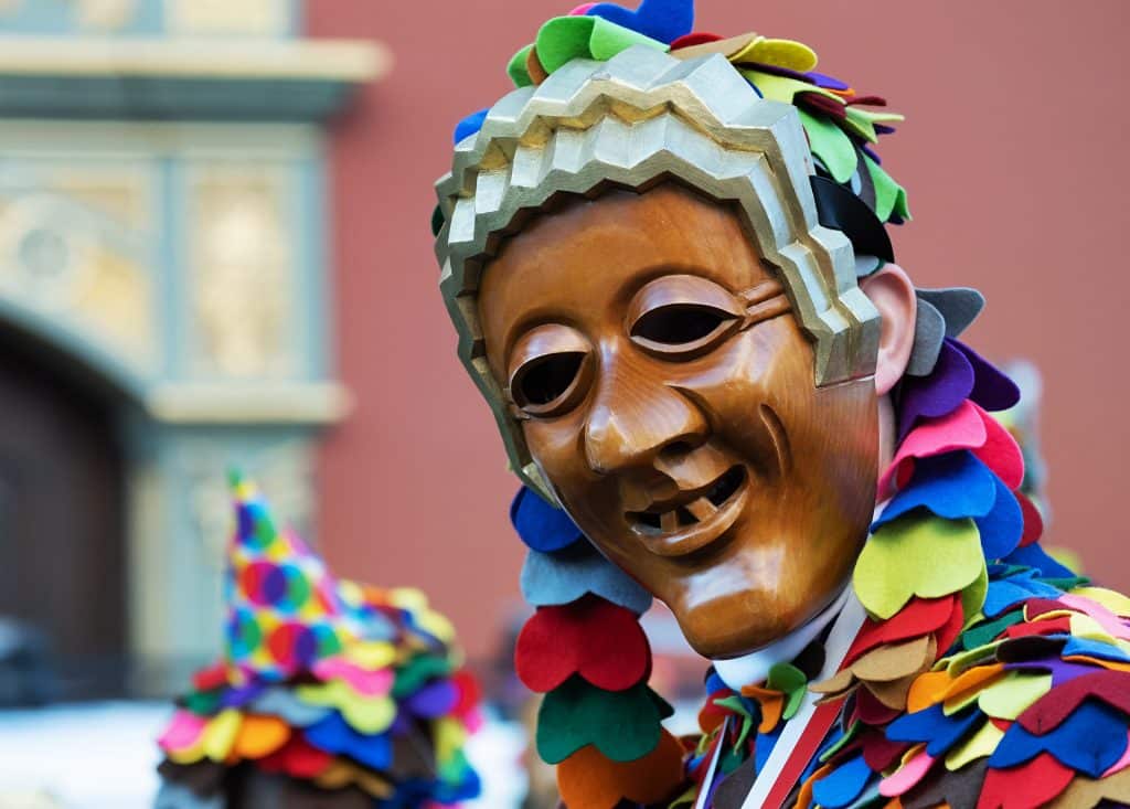 Imagem de uma pessoa fantasiada para o carnaval. A fantasia é colorida e ela usa uma máscara de madeira representando uma pessoa engraçada.