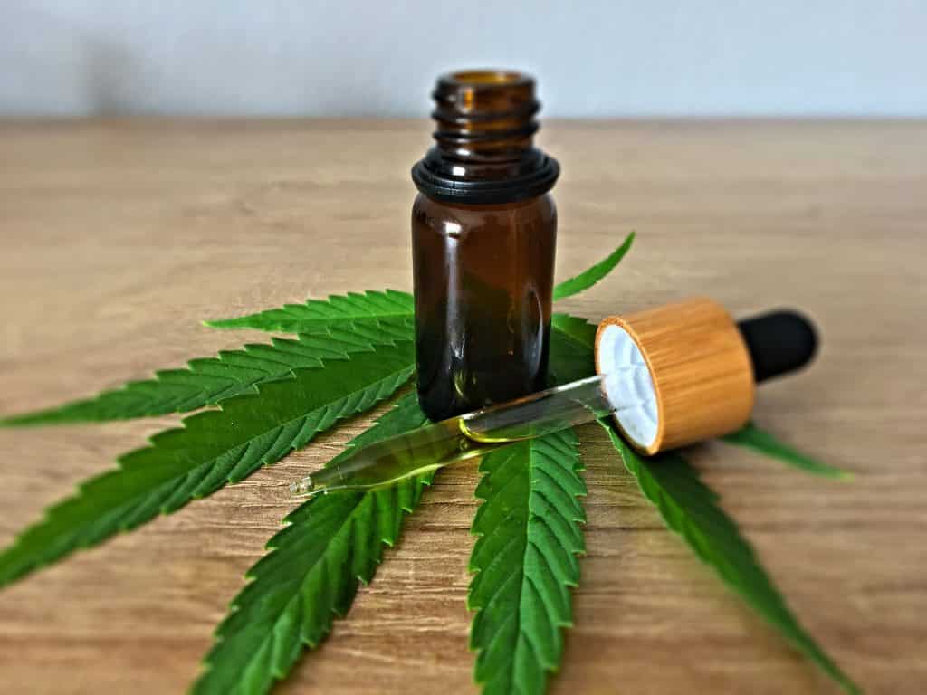 Imagem de uma folha de maconha disposta sobre uma mesa de madeira e sobre a folha um pequeno vidro e um conta gostas com um pouco de óleo da erva dentro dele.