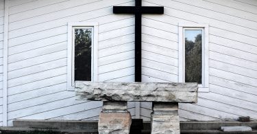 Imagem de fundo branco e em destaque uma cruz na cor preto e o lado dois quadros. À frente um mesa de pedra, representando o altar de uma igreja preparado para a quaresma.