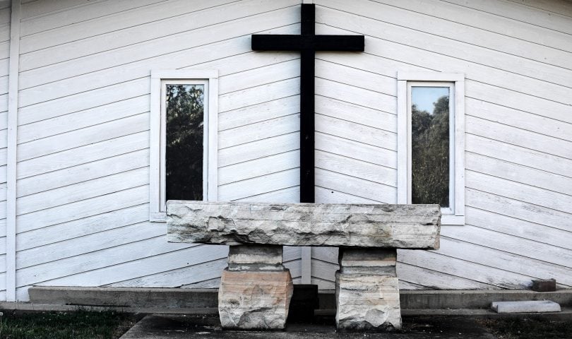 Imagem de fundo branco e em destaque uma cruz na cor preto e o lado dois quadros. À frente um mesa de pedra, representando o altar de uma igreja preparado para a quaresma.