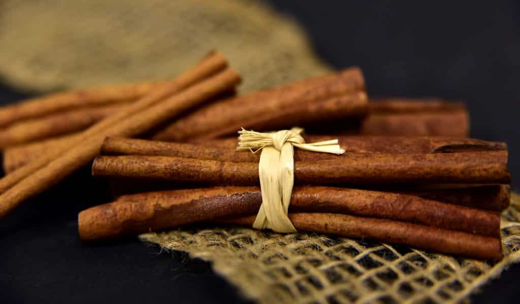 Imagem de vários paus de canela e um pouco delas está arramado com um laço feito com uma tira de palha. Eles estão dispostos sobre uma mesa de fundo preto, decorada com um pedaço de juta.
