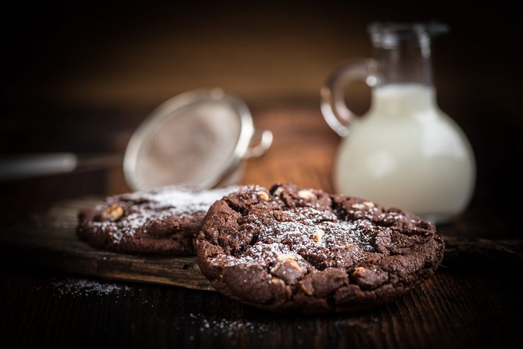 Imagem de cookies de chocolate cobertos com açúcar de confeiteiro, dispostos sobre uma mesa de madeira. Ao lado uma jarra com leite e uma peneira de alumínio.
