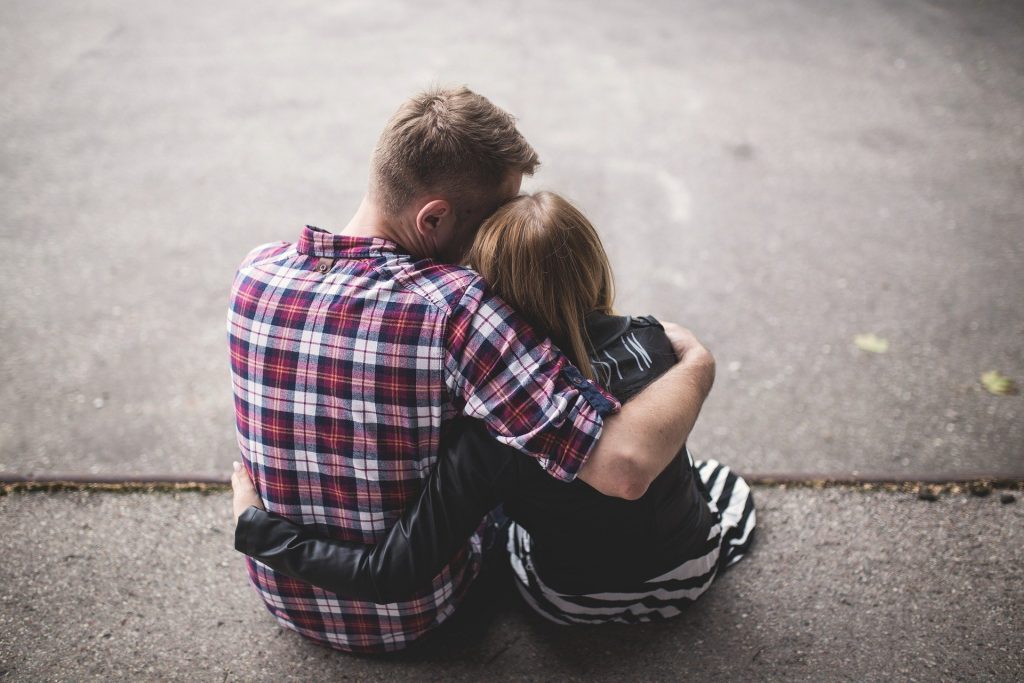 Imagem de um casal abraçados e sentados no chão. Ele usa uma camisa xadrez nas cores vermelho, azul e branco e ela usa um vestido listrado nas cores preto e branco e uma jaqueta na cor preta sobre ele.
