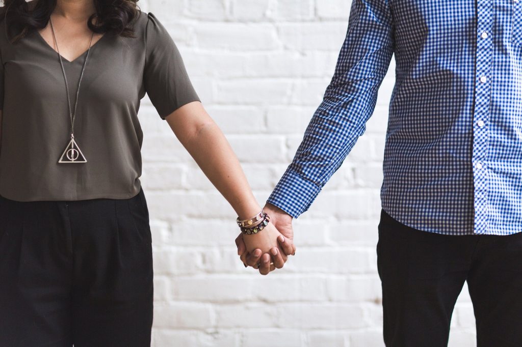 Imagem de fundo branco e em destaque um casal de namorados de mãos dadas. Ela usa uma blusa de manga curta na cor marrom, um colar e uma calça preta. Ele usa uma calça azul escuro e  uma camisa xadrez nas cores azul e branco.