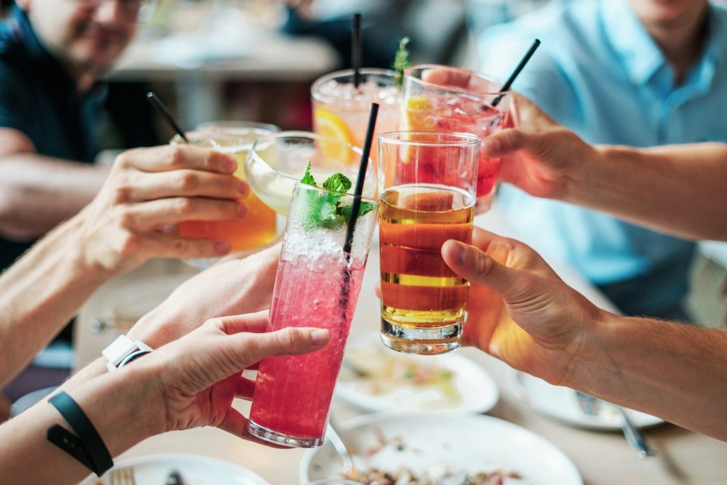 Imagem de várias mãos segurando copos com drinks bem coloridos. Os copos estão decorado e cada um tem um canudo e e alguns possuem uma folha de hortelã.