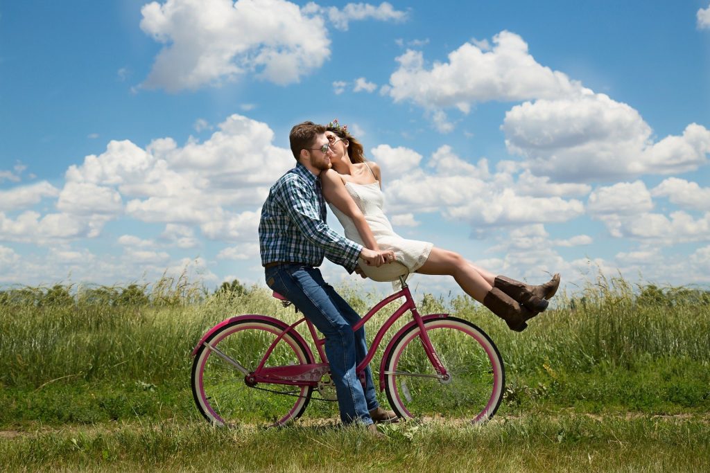Imagem de um casal de homem e mulher. Eles estão em um campo. Ele guia uma bicicleta na cor rosa e a mulher está sentada sobre o guidão da bicicleta. Ele usa calças jeans, camisa xadrez e ela usa um vestido branco, bem casual. Ambos calçam botas na cor marrom.
