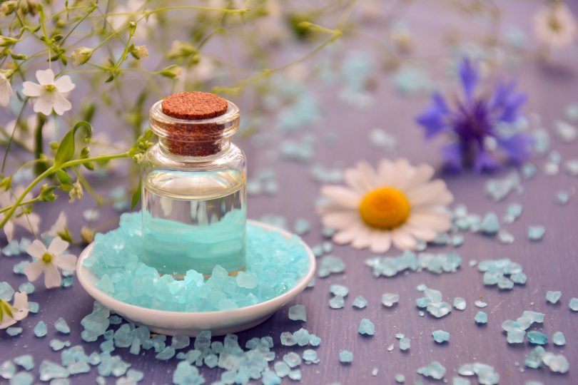 Imagem de um pequeno frasco de vidro contendo óleo essencial. Ele está disposto em um pires branco de porcelana sobre uma mesa decoarada com flores e cristais azul.