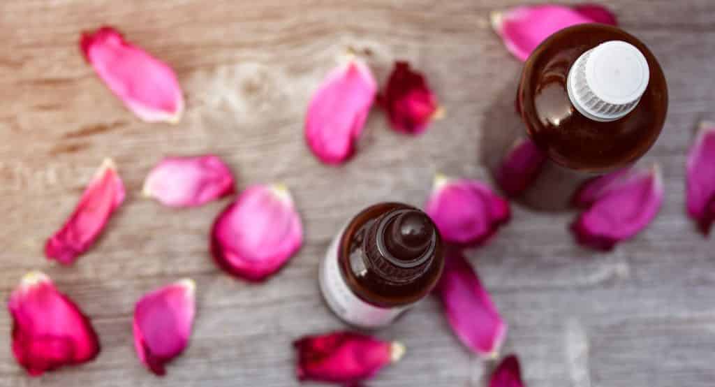 Imagem de dois frascos de vidro marrom contendo óleo essencial. Eles estão dispostos sobre uma mesa de madeira decorada com pétalas de rosas na cor rosa.