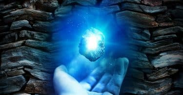Imagem com um fundo de pedras e em destaque a mão de um homem segurando um cristal de água marinha que emana uma luz muito forte na cor azul.
