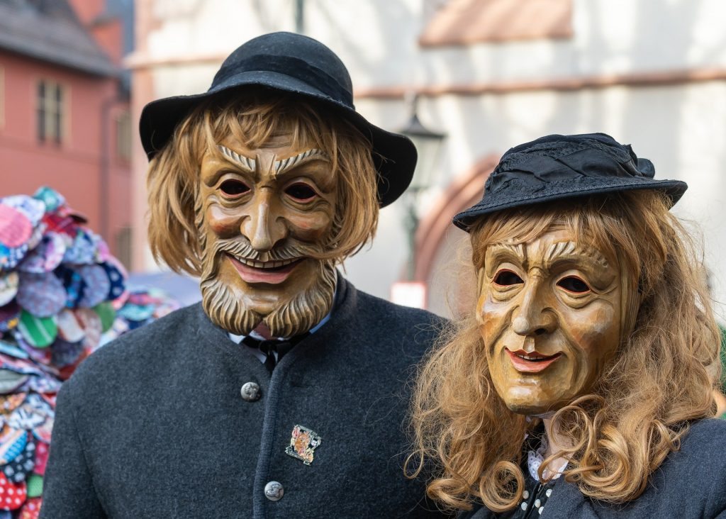 Imagem de um casal, homem e mulher, usando máscara de carnaval de uma série.  Ambos estão na rua, curtindo a festa.