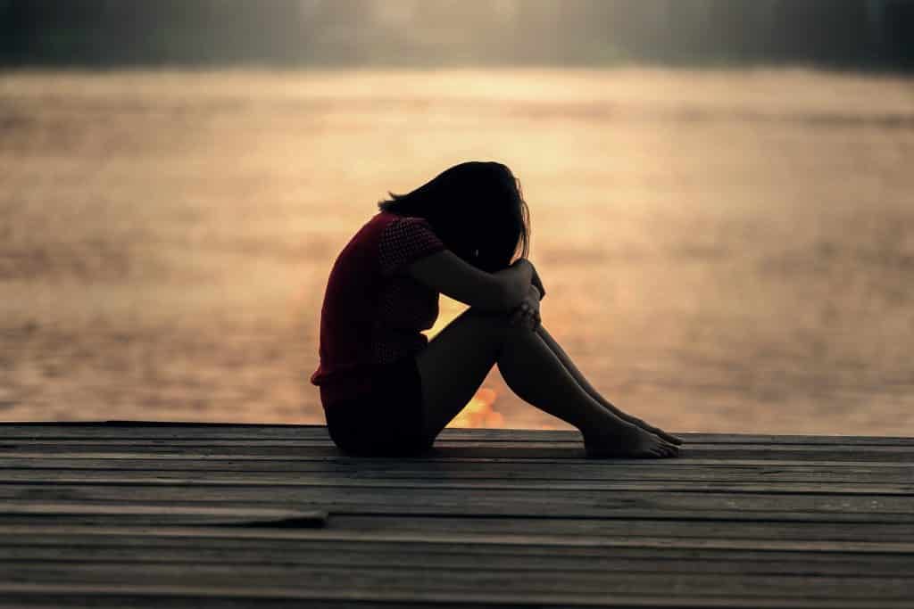 Imagem de uma garota usando shorts e camiseta sentada no chão de um pier e na frente do mar. Ela está triste, com a cabeça baixa.