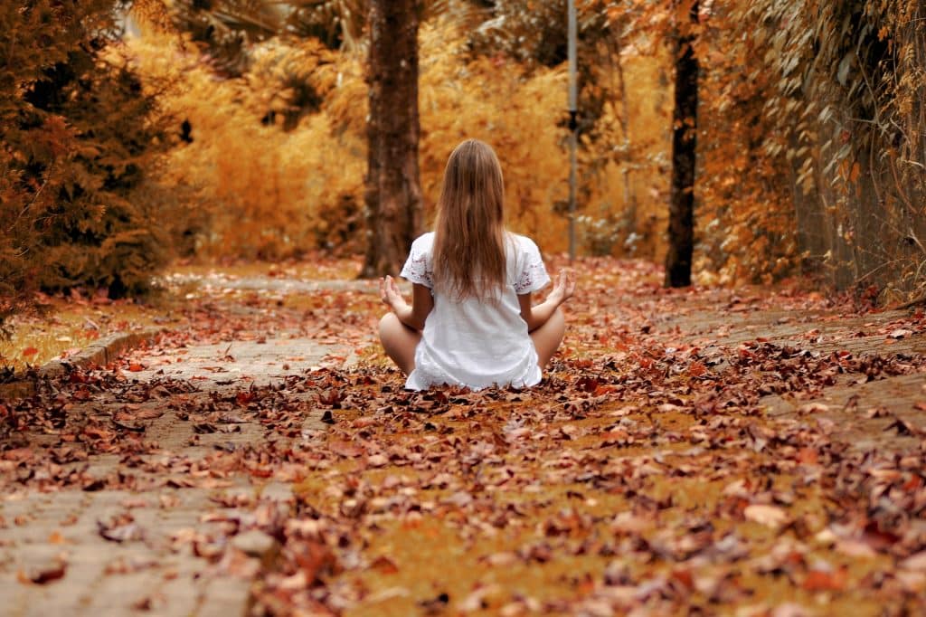 Imagem de uma moça sentada em um chão forrado de folhas. Ela está praticando a meditação.