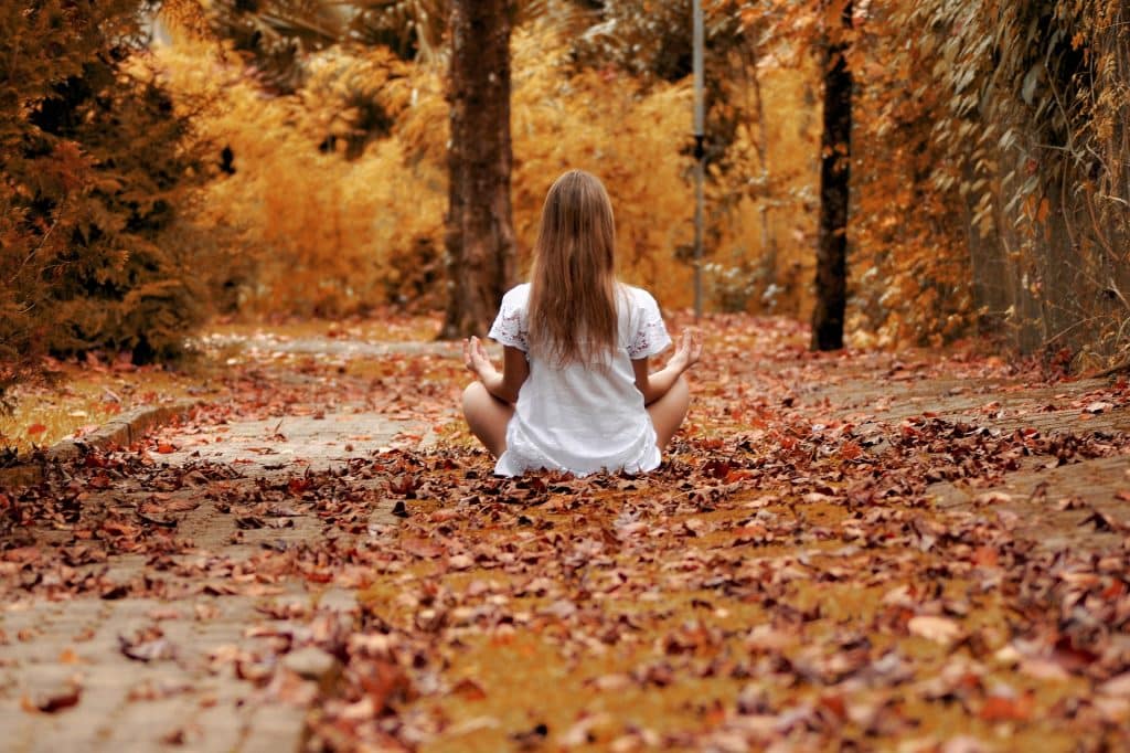 Imagem de uma moça sentada em um chão forrado de folhas. Ela está praticando a meditação.
