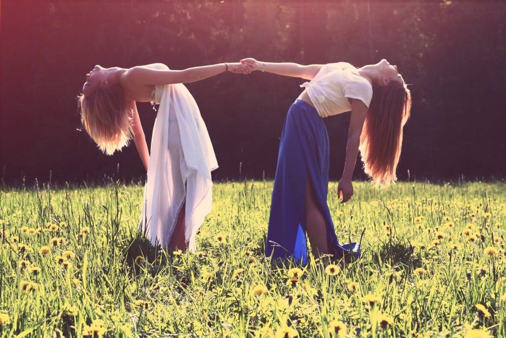 Imagem de duas garotas de mão dadas. Elas estão em um jardim gramado e florida. Ambas usam saias longas, uma na cor azul e a outra na cor branca.