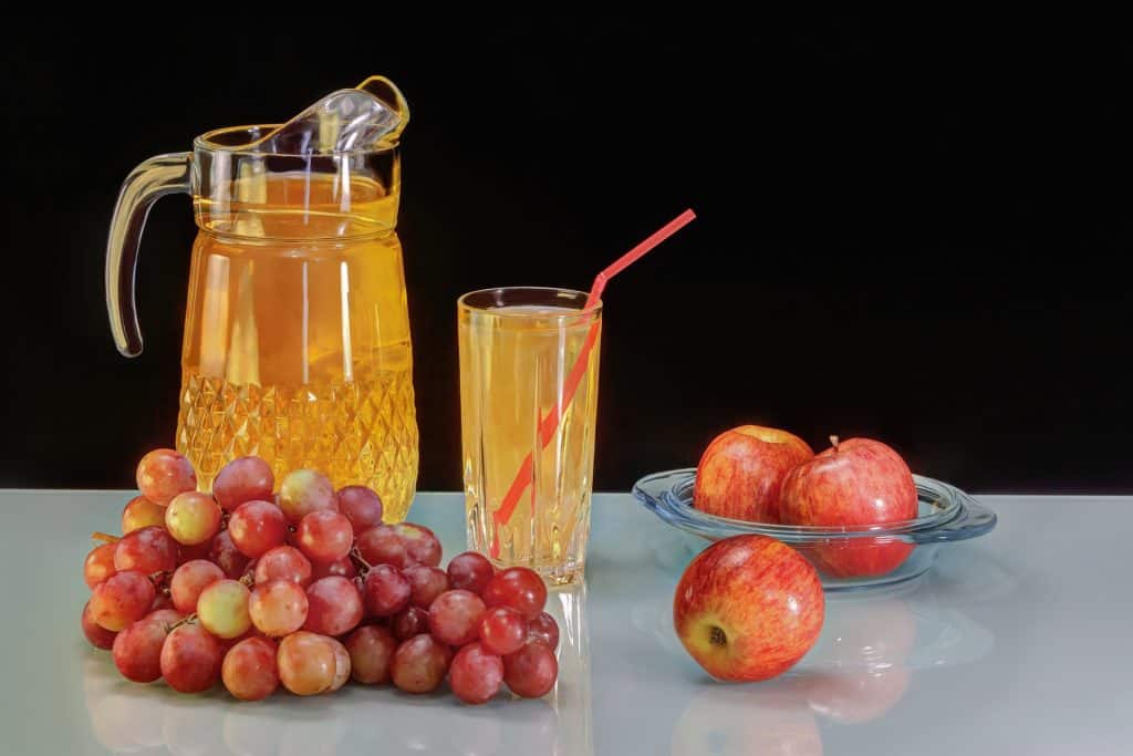 Imagem de fundo preto e em destaque uma jarra cheia de suco de maçã. Ao lado um copo contendo o mesmo suco e um canudo dentro dele. Ao lado e sobre a mesa uma tigela de vidro com duas maçãs vermelhas e ao lado da jarra um cacho de uvas vermelhas e uma maçã.