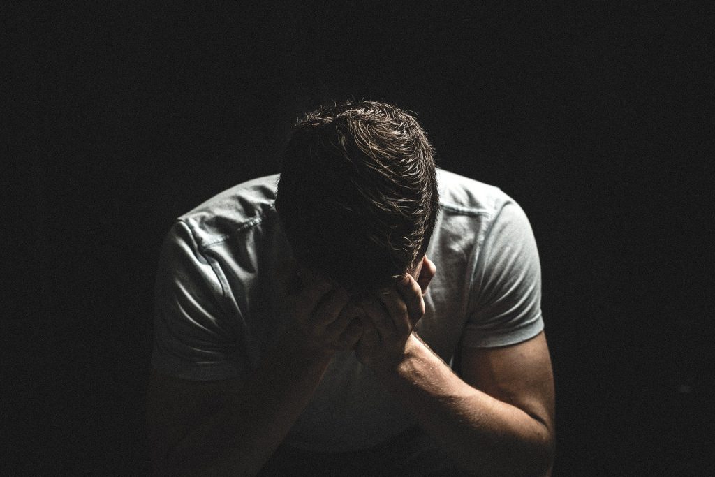 Imagem de fundo preto e nela a figura de um homem de cabeça baixa e com as duas mãos sobre o rosto. Ele está triste.