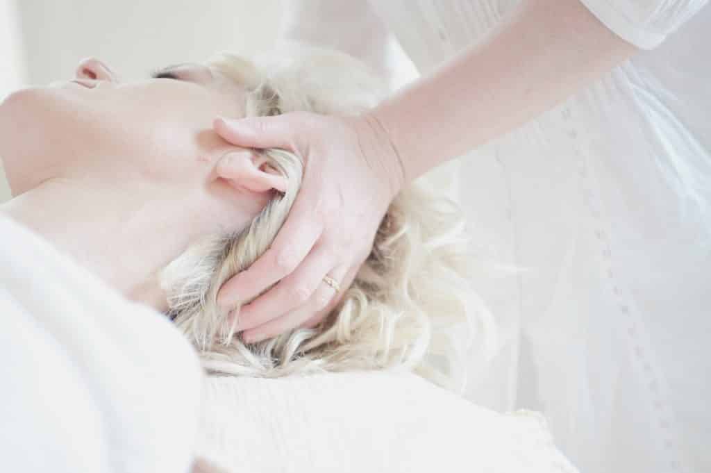 Imagem de uma mulher loira de cabelo curtos, deitada sobre uma maca. Em pé temos uma mulher que usa um camisão branco e está impondo levemente uma de suas mãos sobre a cabeça da paciente.