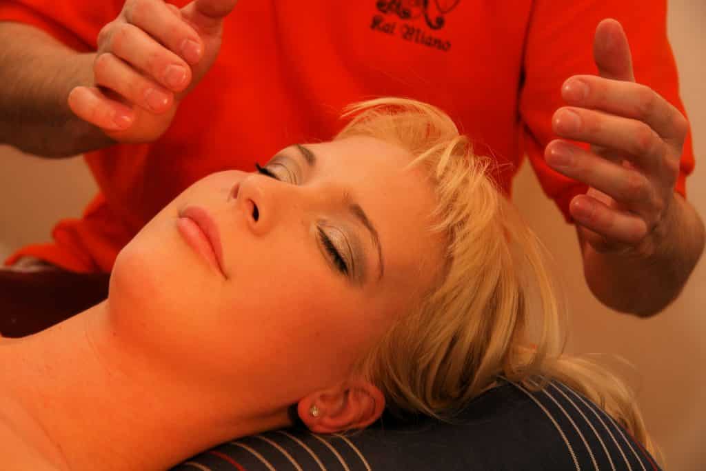 Imagem de uma mulher loira deitada sobre uma maca. Ela está recebendo reiki de um homem que está posicionando suas mãos sobre a cabeça dela. Ele usa camiseta na cor laranja.