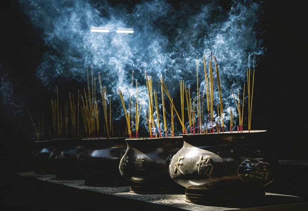 Imagem de cinco panelas com símbolos chineses desenhados, dentro delas vários incensos de citronelas sendo queimados e saindo muita fumaça.
