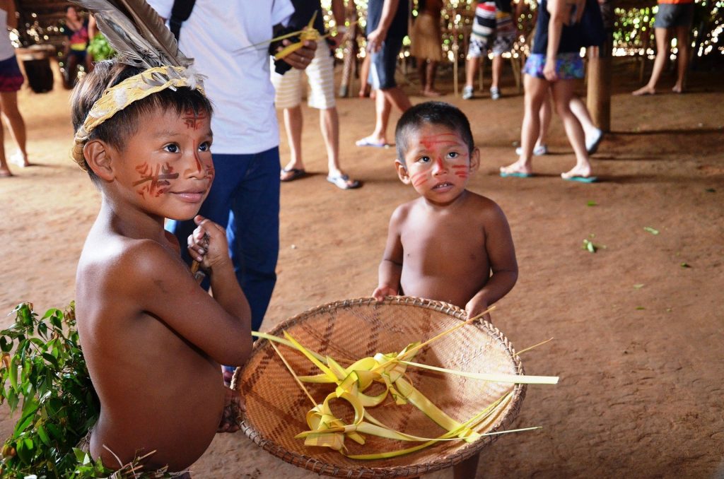 Imagem de duas crianças indígenas segurando um cesto de vime. Elas estão com os rostos pintados e estão dentro do terreiro na tribo.
