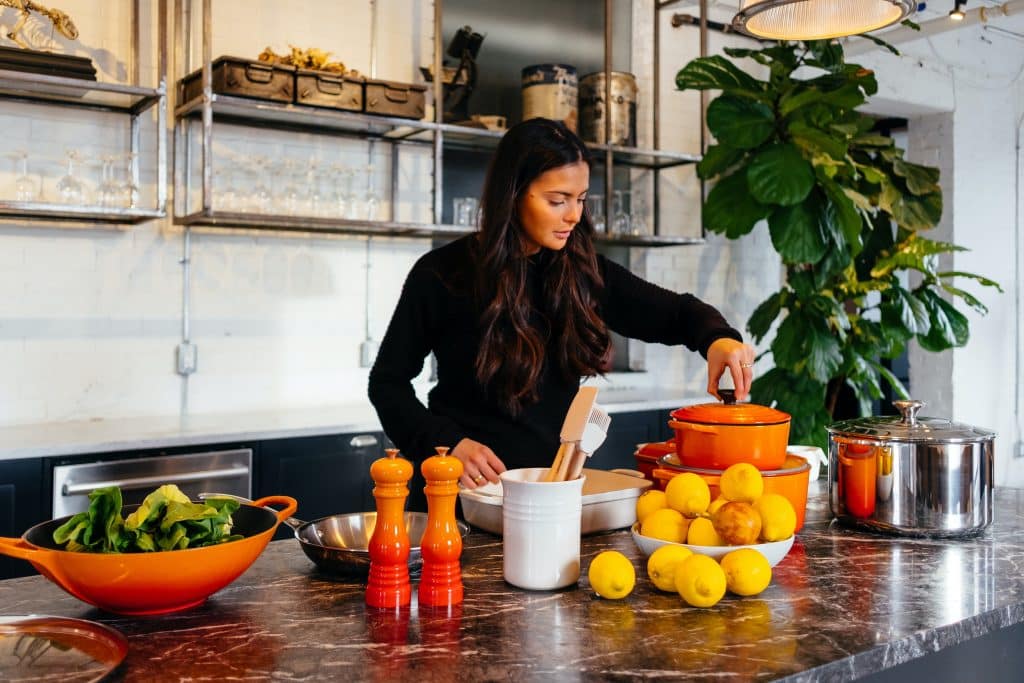 Imagem de uma cozinha e uma linda bancada de mármore. Em destaque uma mulher de cabelos longos e blusa de frio na cor preta. Ela está cozinhando. Sobre a bancada temos vários tipos e modelos de panelas na cor laranja e alguns ingredientes como verdura e limões.
