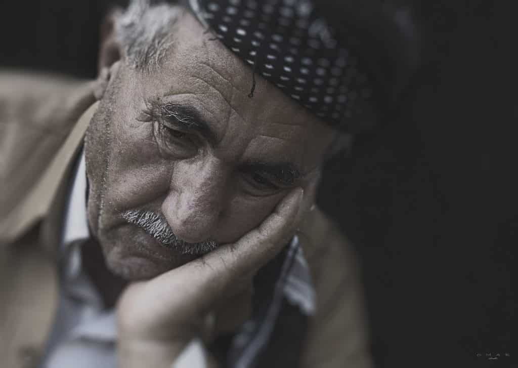 Imagem de um senhor triste, cabisbaixo. Ele está com uma de suas mãos segurando um do lado do seu rosto. Ele usa bigodes e uma boina sobre a cabeça.
