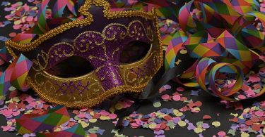 Imagem de uma máscara de carnaval nas cores dourada e roxa. Ela está sobre uma mesa de fundo preto e na mesa estãoo espalhados confetes e serpentinas coloridas.