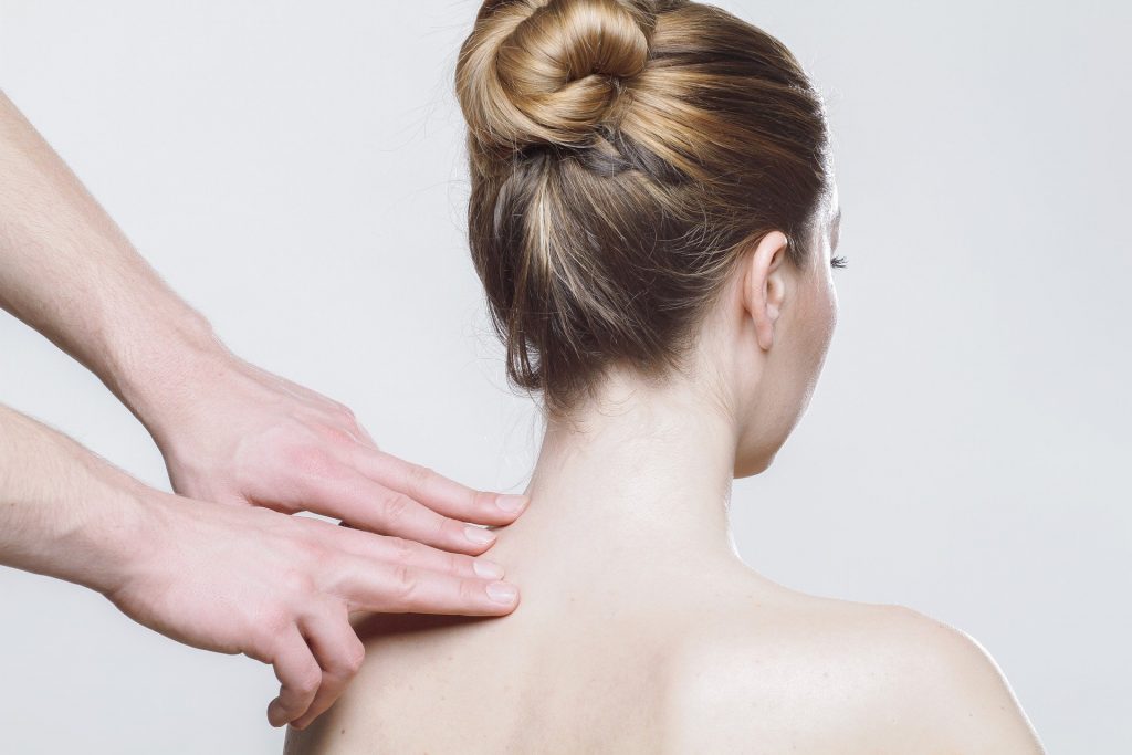 Imagem de uma mulher de cabelo preso, sentada de costas. Em pé um homem que está colocando suas mãos sobre um lado do ombro da paciente.