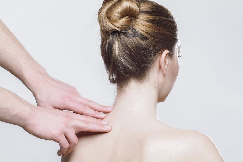 Imagem de uma mulher de cabelo preso, sentada de costas. Em pé um homem que está colocando suas mãos sobre um lado do ombro da paciente.
