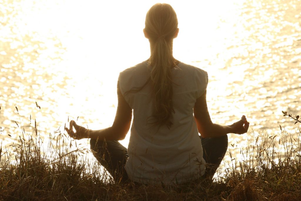 Imagem de uma mulher loira de costas praticando a meditação em frente de um rio. Ela está sentada em um gramado. Usa uma calça escura e uma camiseta branca.