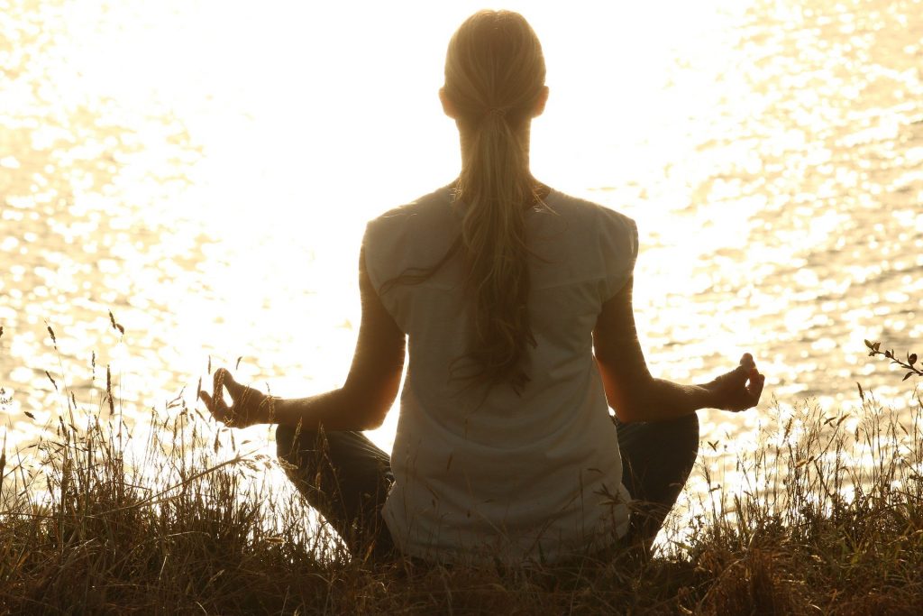 Imagem de uma mulher loira de costas praticando a meditação em frente de um rio. Ela está sentada em um gramado. Usa uma calça escura e uma camiseta branca.
