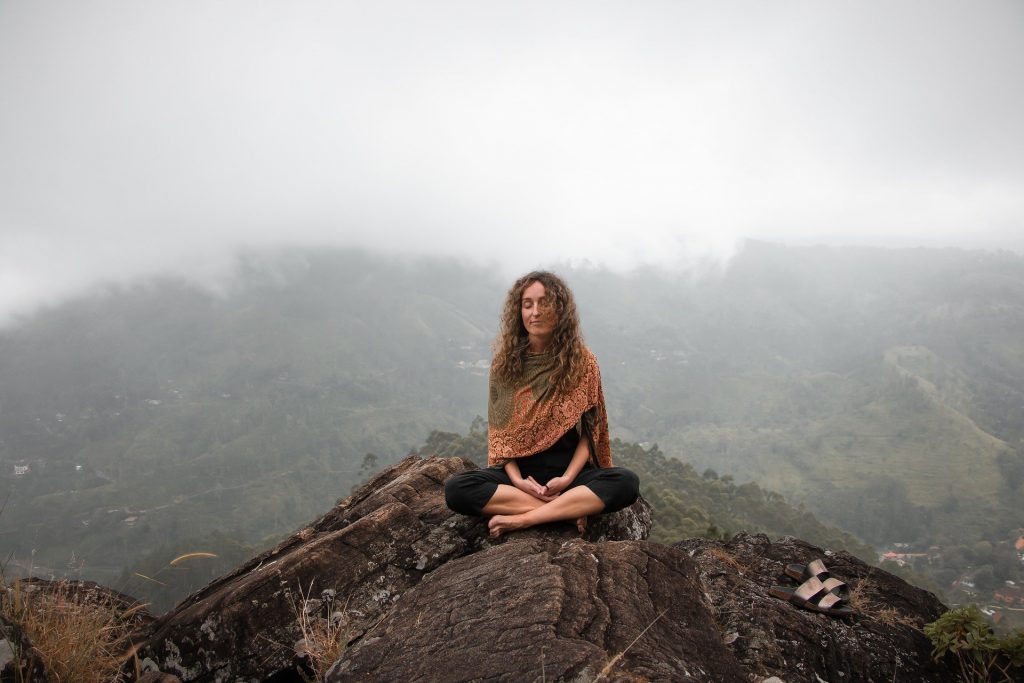 Imagem de uma montanha com neblina. Em destaque uma mulher meditando sobre uma rocha.