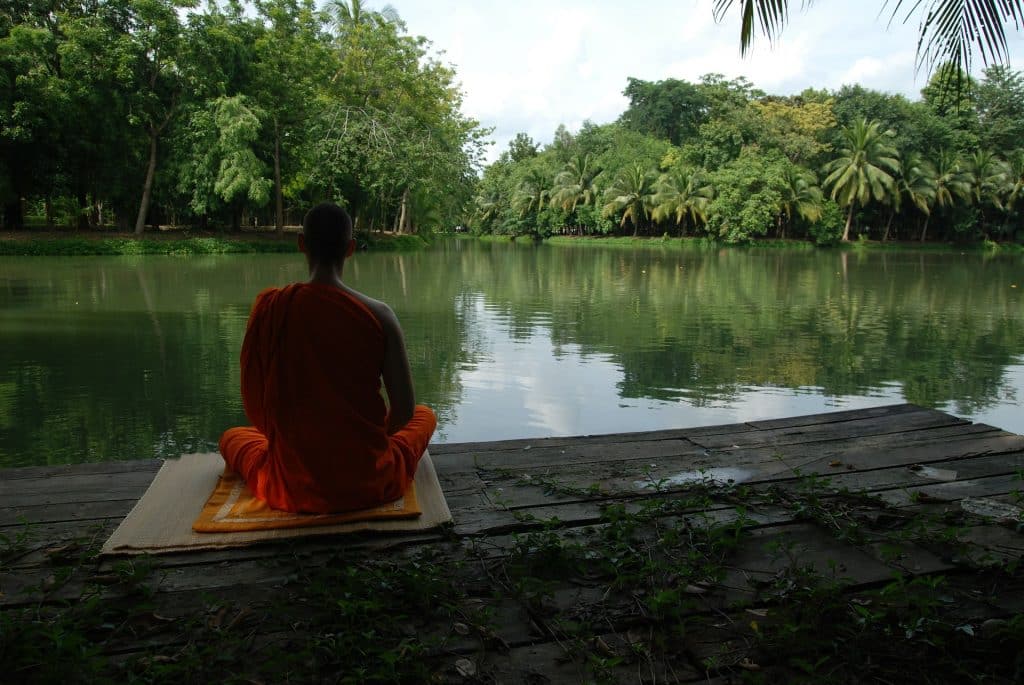 Imagem de um homem sentado de costas sobre um tapete de yoga praticando a meditação em frente a um lago todo arborizado.
