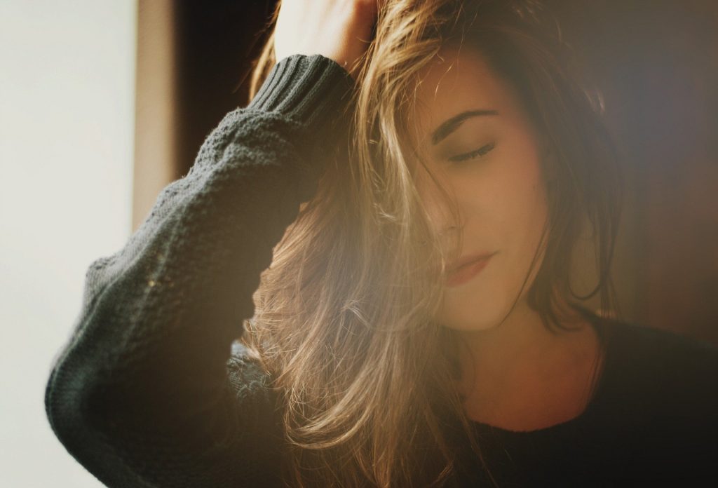 Imagem de uma mulher próxima à janela com uma luz refletindo os seus cabelos castanhos claros. Ela usa uma blusa de lã na cor preta.
