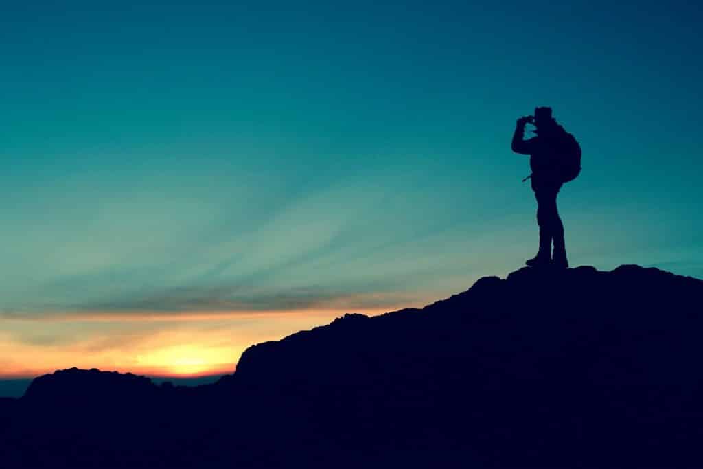 Imagem de um homem no topo de uma montanha. Ele está com uma mochila nas costas e usa um binóculo para observar o lindo pôr do sol no final da imagem.