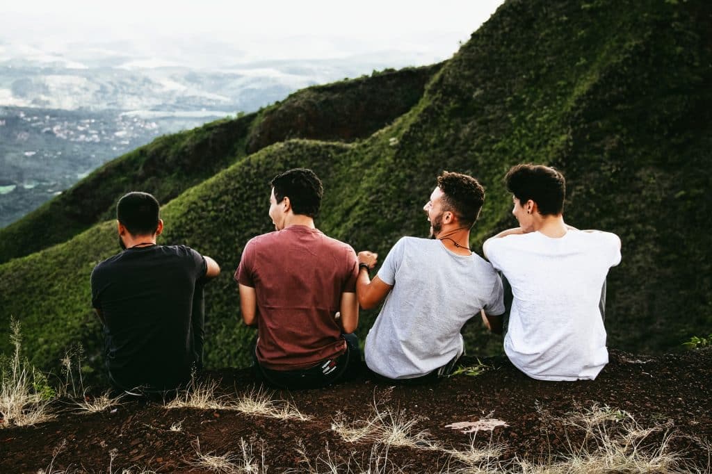 Imagem de quatro rapazes sentados em uma montanha apreciando a natureza. Todos usam camiseta de malha, nas cores preto, vinho, cinza e branco.
