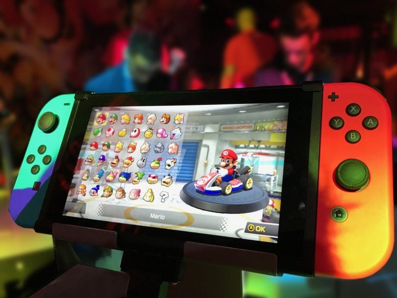 Imagem do controle remoto de um videogame. Na tela do controle imagens de vários ícones de jogos e ao fundo a imagem do Super Mario Bros.