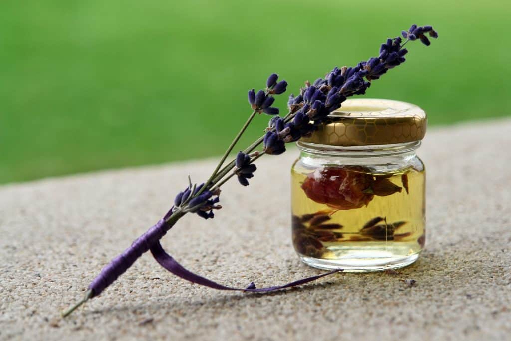 Imagem de um pote de vidro transparente contendo óleo essencial de lavanda. Sobre ele um ramo de lavanda decorado com uma fita na cor roxa.
