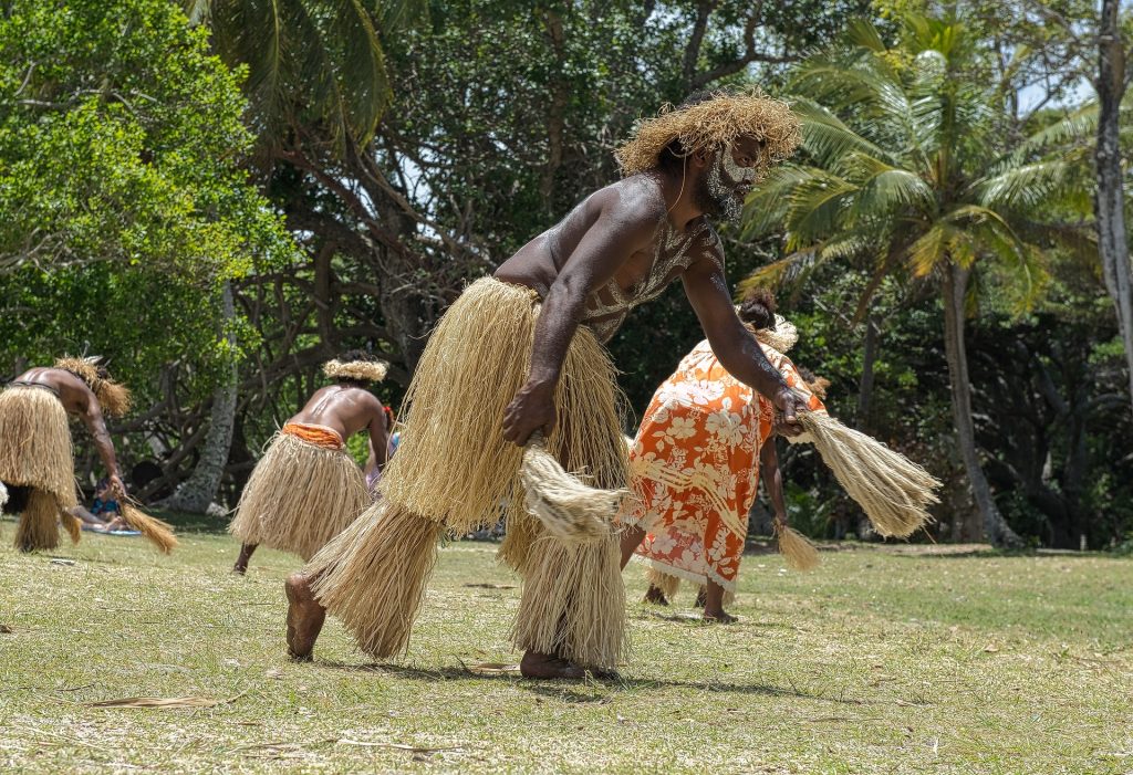 Imagem de homens e mulheres indígenas dançando no terreiro gramado da tribo. Eles usam saias feitas e barbante com um adereço do mesmo material na cabeça. Elas usam vestido longo na cor laranja com flores brancas.

