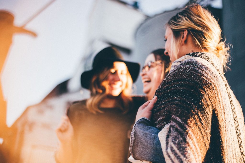 Imagem de fundo iluminado e em destaque, três mulheres sorridentes. Uma usa um chapéu preto, outra um óculos de grau e a outra o cabelo preso. Todas estão vestidas com roupas de frio.