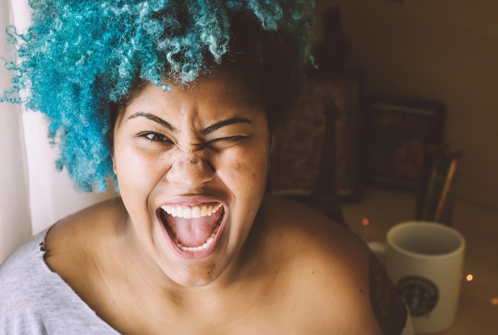 Imagem de uma mulher morena super sorridente e feliz. Ela tem uma parte do cabelo pintado na cor azul. Usa camiseta cinza de malha e possui uma tattoo no braço direito da tela.