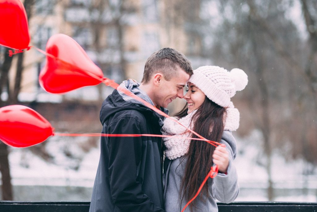 Imagem de um casal um olhando de frente para o outro. Ambos estão agasalhados. Ela usa uma rouca de lã e um cachecol também de lã. Ela segura três balões vermelhos em formato de coração.