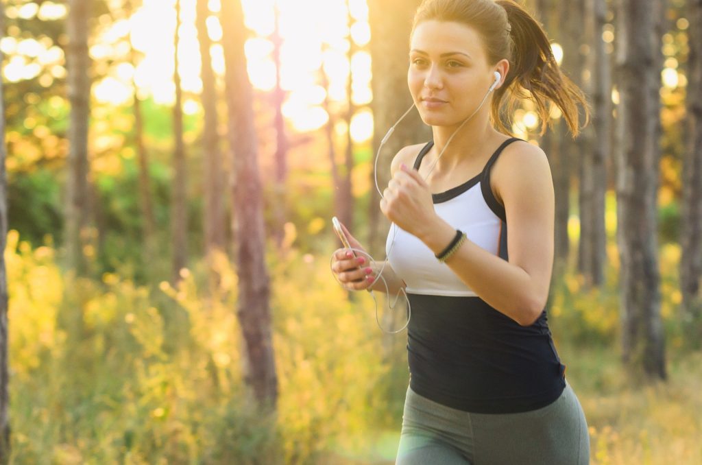 Imagem de uma jovem mulher fazendo exercício físico, ou seja, ela está correndo em um lugar arborizado. Ela usa roupa esportiva e houve música do seu celular com um fone de ouvido.
